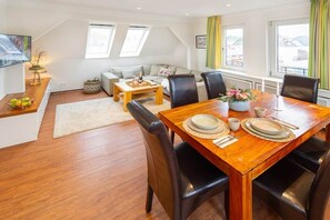 A dining area with a wooden table set for four, a sofa, and a coffee table in a bright room with skylights. - To complete (Wangerooge)