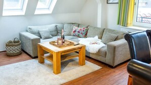 A living room with a sofa, a wooden coffee table, a window with curtains, and a framed picture on the wall. - To complete (Wangerooge)