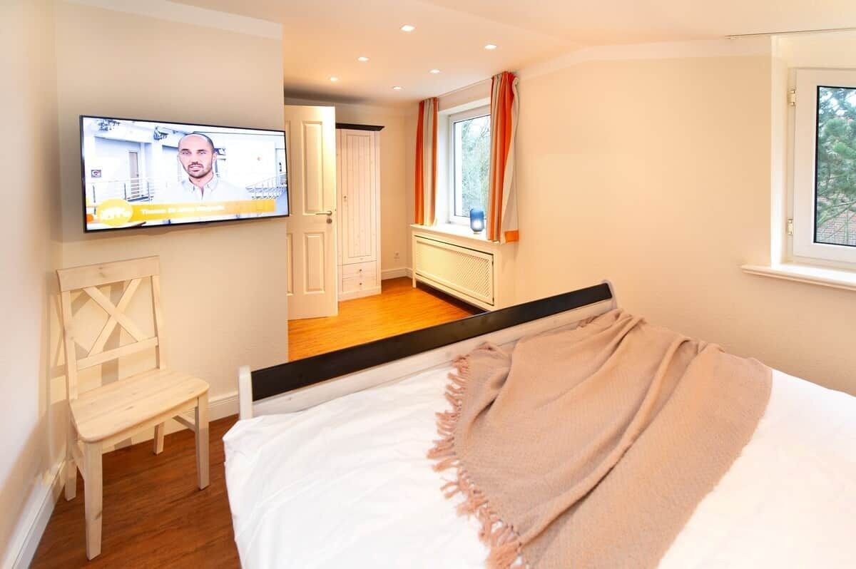 Quarto com cama, televisão na parede, janela com cortinas e uma cadeira.