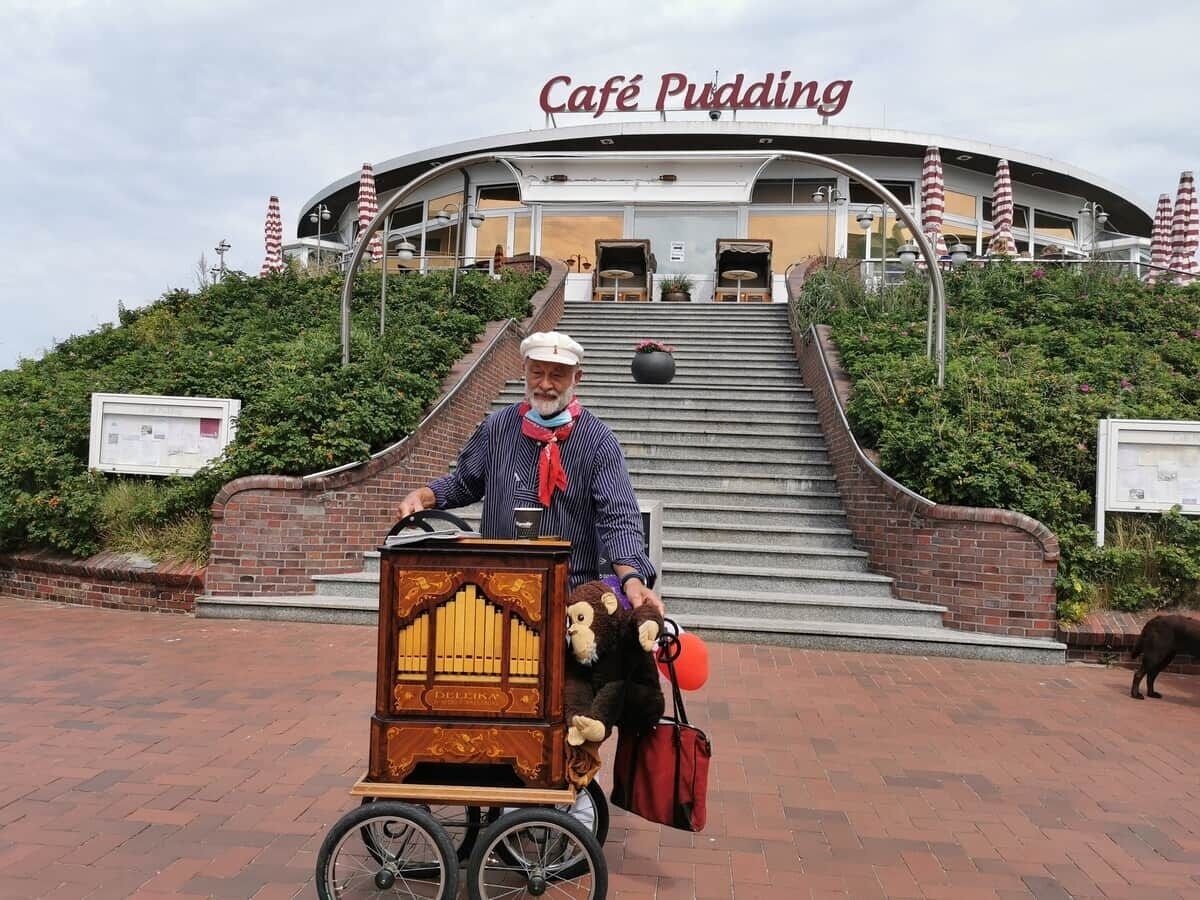 Una persona en bicicleta jala un carrito con una caja de música, frente a un edificio con un letrero que dice 'Café Pudding'.