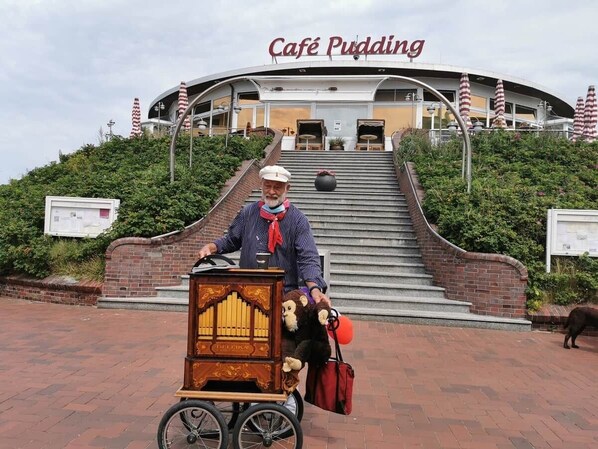A person on a bicycle with a music box cart, in front of a building with a sign reading 'Café Pudding'.
