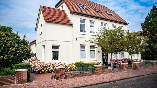 A two-story residential building with a red-tiled roof, white walls, and a brick pathway leading to the entrance. - Feuerstein Family Home, Apartment 5 (Wangerooge)