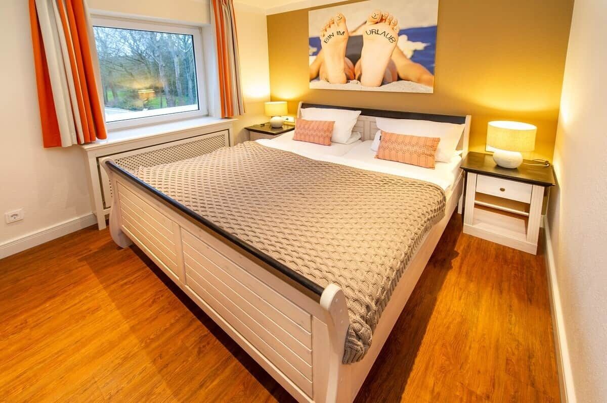 Un dormitorio con cama, mesita de noche, lámpara, ventana con cortinas y un cuadro en la pared.