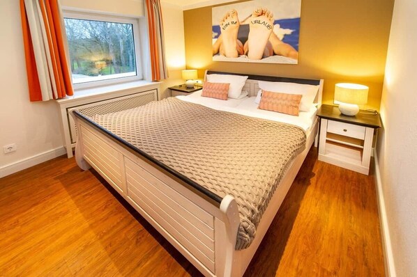 A bedroom with a bed, a nightstand, a lamp, a window with curtains, and a painting on the wall.