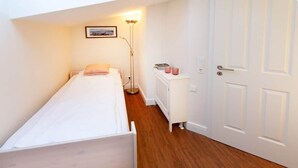 A small, single-bed room with a wooden floor, a standing floor lamp, a radiator, and a white door. - Feuerstein Family Home, Apartment 5 (Wangerooge)