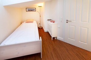 A small, single-bed room with a wooden floor, a standing floor lamp, a radiator, and a white door.