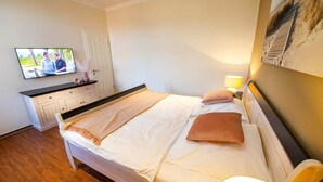 Hotellihuone, jossa on sänky, seinälle asennettu televisio, yöpöytä lampun kanssa ja taulu seinällä.