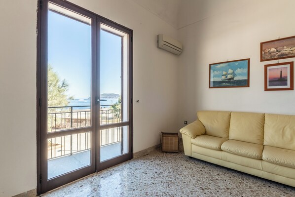 A room with a large sliding glass door leading to a balcony, a beige sofa, and two framed pictures on the wall. - La Finestra sul Blu (Favignana)