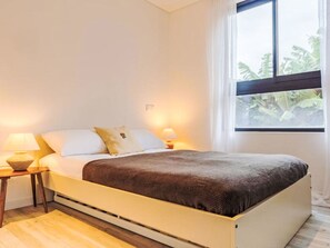 Ein Bett mit Kopfteil, zwei Nachttische mit Lampen, ein Fenster mit Blick auf Grünflächen und eine helle Wand.