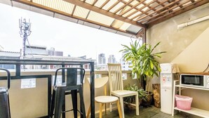 Ein Balkon mit Blick auf die Stadt, eine Mikrowelle, ein Stuhl, ein Blumentopf und ein Mülleimer.