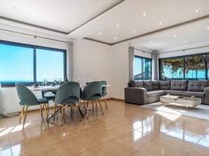 Ein geräumiger Wohnbereich mit Esstisch, Stühlen, einer Couch und einem Couchtisch. Große Fenster bieten einen Ausblick ins Freie.