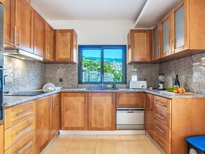 A kitchen with wooden cabinets, granite countertops, a built-in dishwasher, and a window with a view of a coastal landscape. - Camara de Lobos Lux by Interhome (Câmara de Lobos (Madeira))