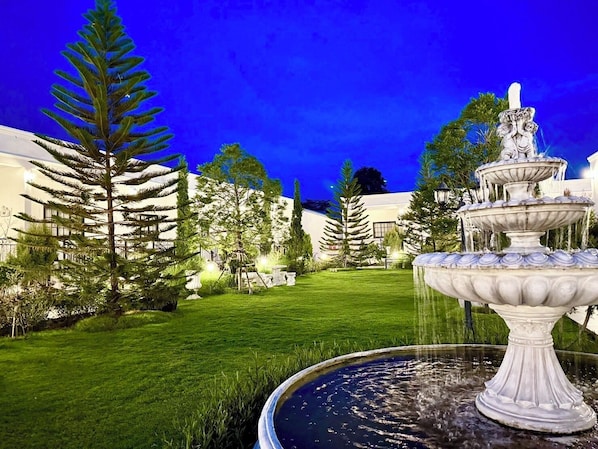 A nighttime view of a garden with a multi-tiered fountain, well-manicured grass, and a building with illuminated windows in the background. - Inter Paradise Resort (Dan Khun Thot)