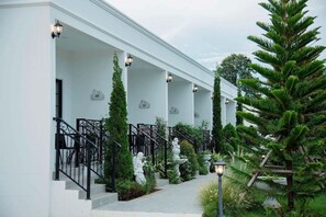 Ein weißes Gebäude mit nummerierten Balkonen, einer Steinstatue und einem gepflegten Garten mit einem großen Baum.