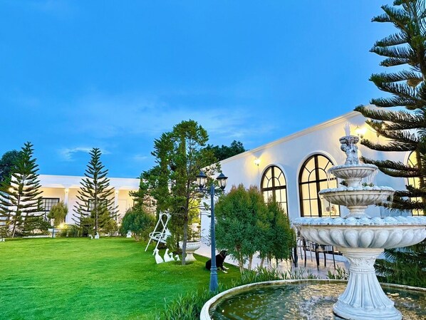 A well-maintained garden with a fountain, a dog walking, and a building with arched windows. - Inter Paradise Resort (Dan Khun Thot)