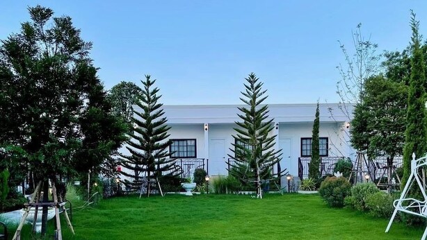 A grassy outdoor area with a white building in the background, surrounded by trees and a swing set. - Inter Paradise Resort (Dan Khun Thot)