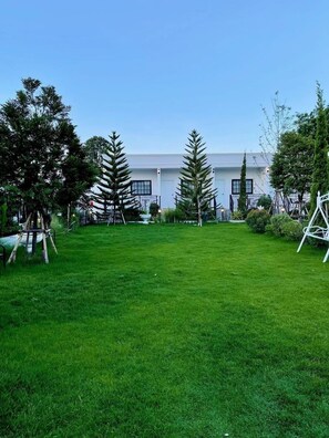 A grassy outdoor area with a white building in the background, surrounded by trees and a swing set. - Inter Paradise Resort (Dan Khun Thot)