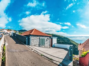 Eine Küstenstraße mit einem Steinhaus mit rotem Dach, einer gepflasterten Straße und Blick auf das Meer und die Hügel im Hintergrund.