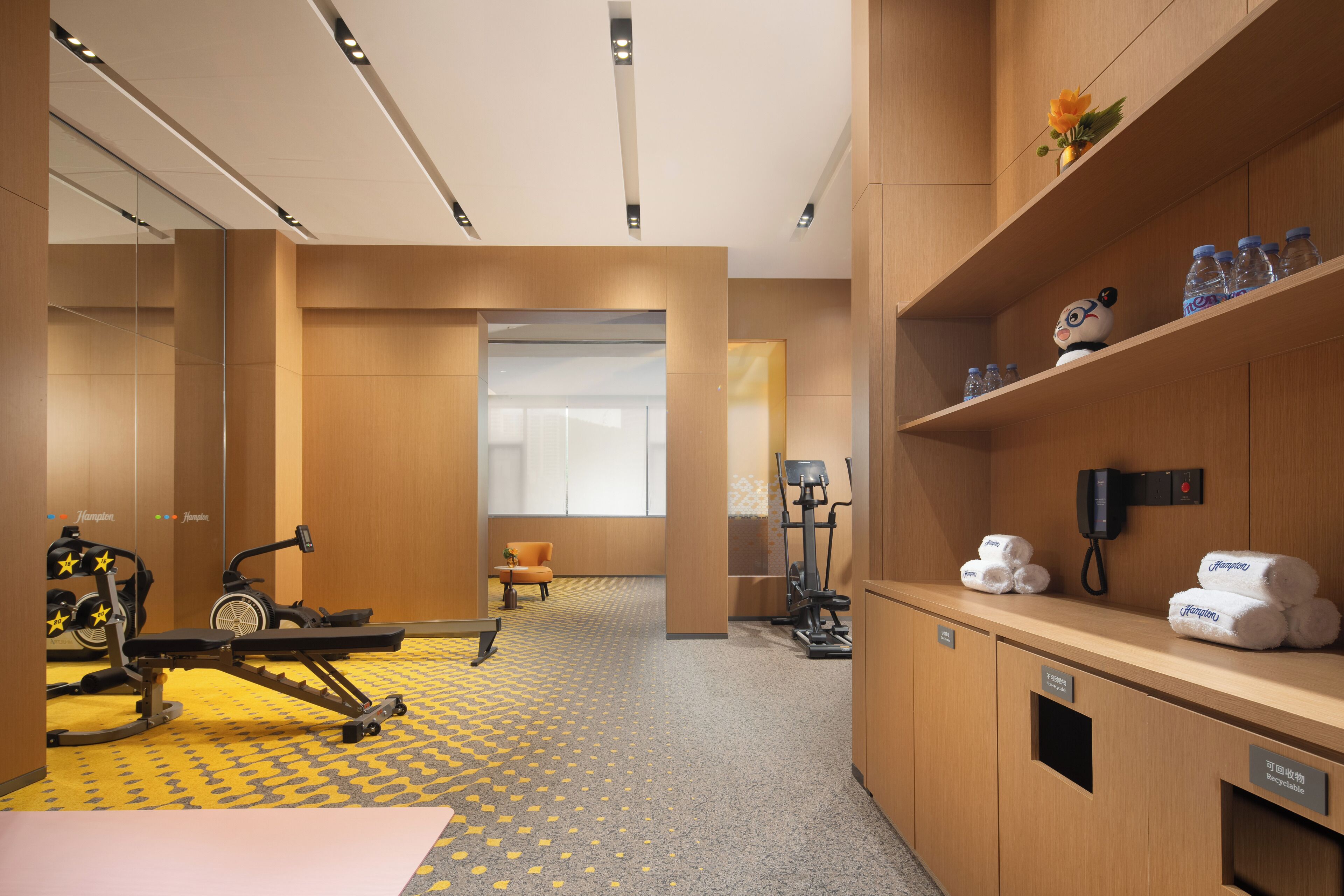 Habitación de hotel con zona de ejercicio, equipo para entrenar, sofá y estante con toallas y botellas.
