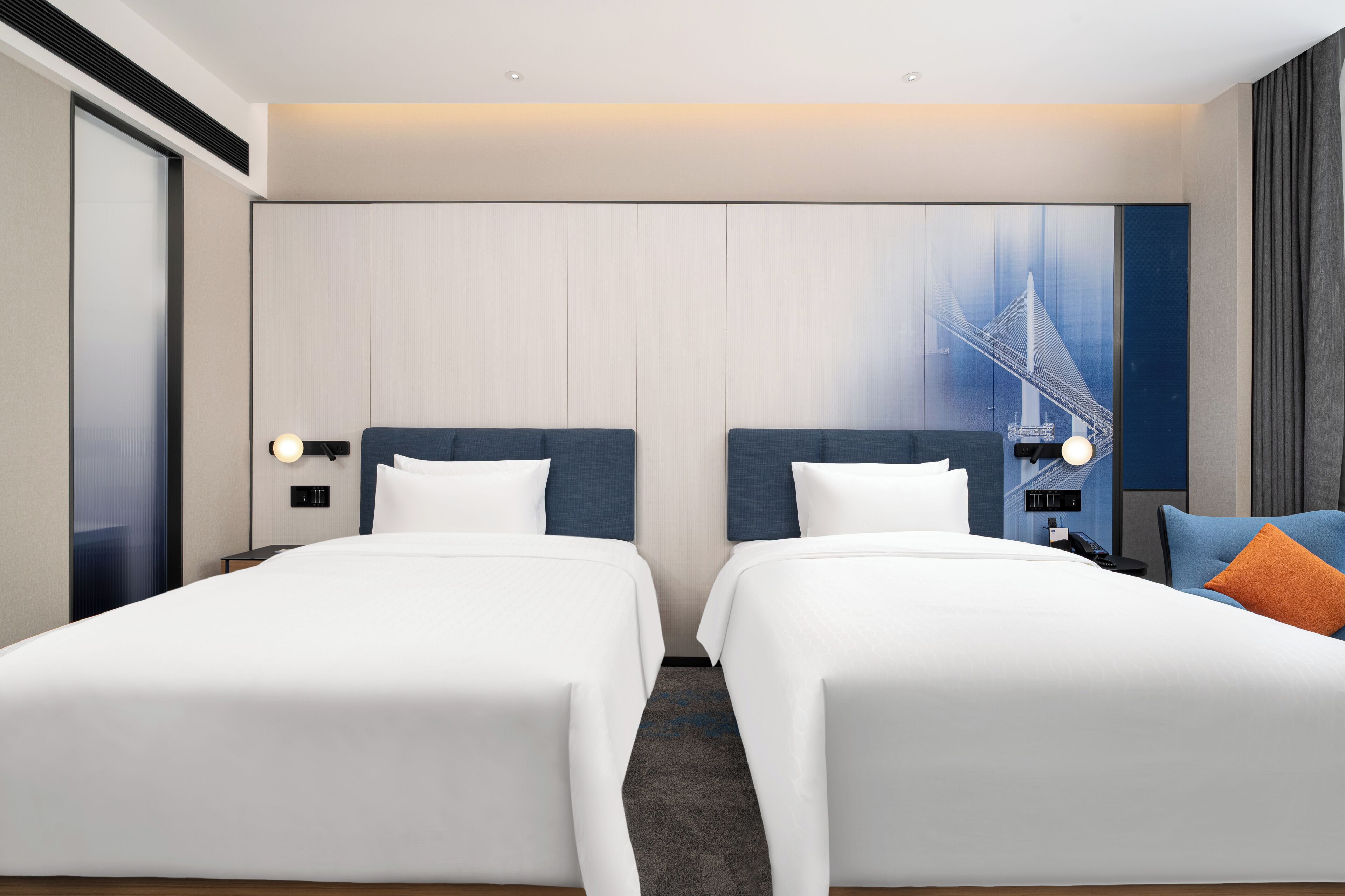 A hotel room with two beds, a large window with a cityscape view, a wall-mounted phone, and a nightstand with a lamp.