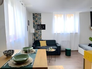 A modern living room with a sofa, a dining table with bowls and cutlery, a small side table, and a large window with curtains. - 'F1 Fully Equipped Apartment Near Tram - Jungle' with Wi-Fi and Air Conditioning (Mulhouse)