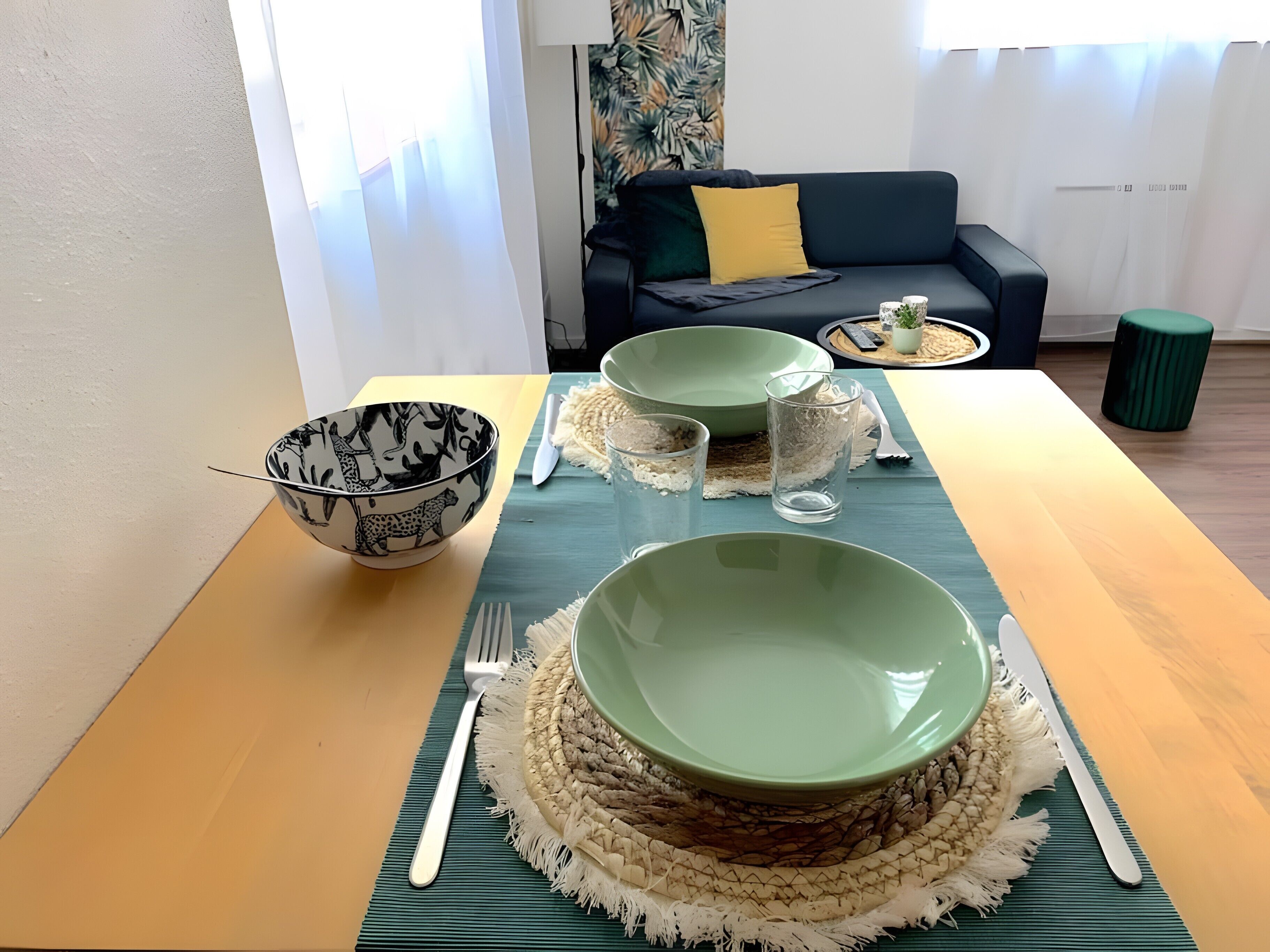 Een eettafel gedekt met groene kommen, een zwart gebekeerde kom en bestek, met een bank en gordijnen op de achtergrond.