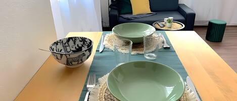 Een eettafel gedekt met groene kommen, een zwart gebekeerde kom en bestek, met een bank en gordijnen op de achtergrond.