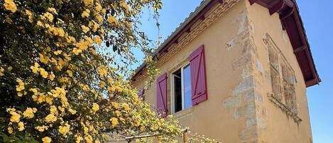 A stone building with a red roof, pink shutters, and a metal gate.