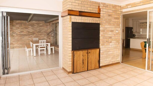 A room with a brick fireplace, a dining table with chairs, and a kitchen area visible through an open door. - Charming 4-bedroom Apartment in Jeffreys Bay, South Africa (Jeffreys Bay)