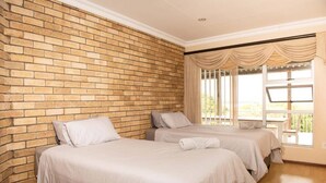 A room with two beds, a brick wall, a window with curtains, and a balcony. - Charming 4-bedroom Apartment in Jeffreys Bay, South Africa (Jeffreys Bay)