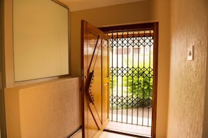 A wooden door with a metal gate, a view of greenery outside, and a light switch on the wall. - Charming 4-bedroom Apartment in Jeffreys Bay, South Africa (Jeffreys Bay)