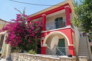 A building with a red facade, a balcony with green shutters, and a stone wall with a bush.