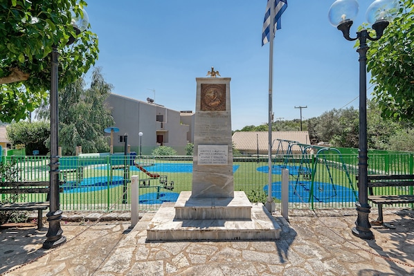 A monument with inscriptions, surrounded by a playground with a green fence, and a building in the background.