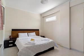 A neatly made bed with white bedding and a folded towel, a bedside table with a lamp, and a window with a view of the outside.