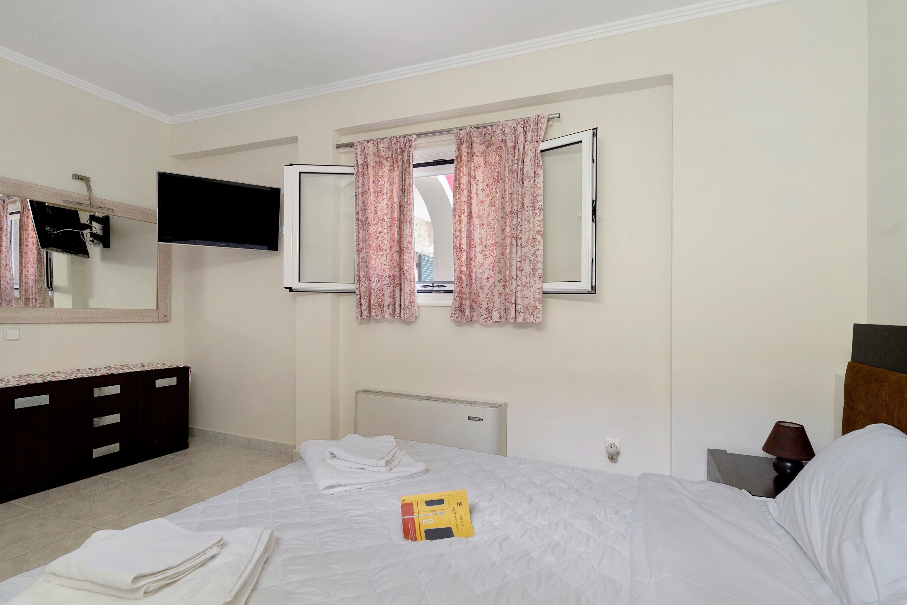 Una camera d'albergo con un letto, una TV a schermo piatto fissata al muro, una finestra con tende floreali, un comodino con una lampada e un comò di colore scuro.