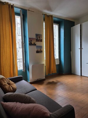 A room with a sofa, a radiator, a door, and a window with curtains. - CHICKY HOUSE (Paris)