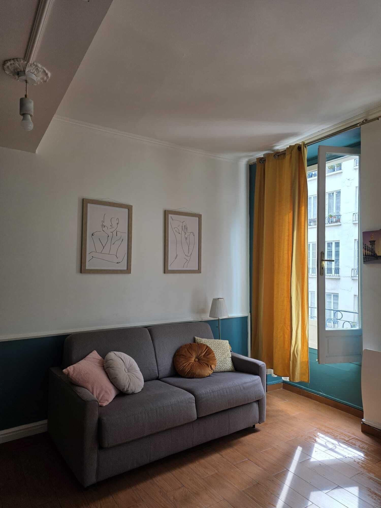Un soggiorno con un divano grigio, due opere d'arte incorniciate, una lampada e una vista attraverso una porta con tende gialle.
