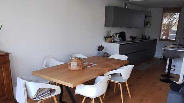 Ett modernt vardagsrum med ett matbord i trä, vita stolar, en grå soffa och ett kök i bakgrunden.