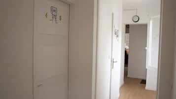 Un couloir avec des portes blanches, dont l’une porte un numéro et un symbole indiquant la présence de toilettes. Il y a une horloge au mur et une porte qui donne sur une autre pièce.