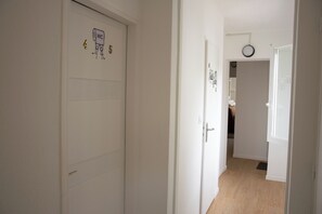Ein Flur mit weißen Türen, eine davon ist mit einer Nummer und einem Toilettensymbol gekennzeichnet. An der Wand hängt eine Uhr und eine Tür führt in einen weiteren Raum.