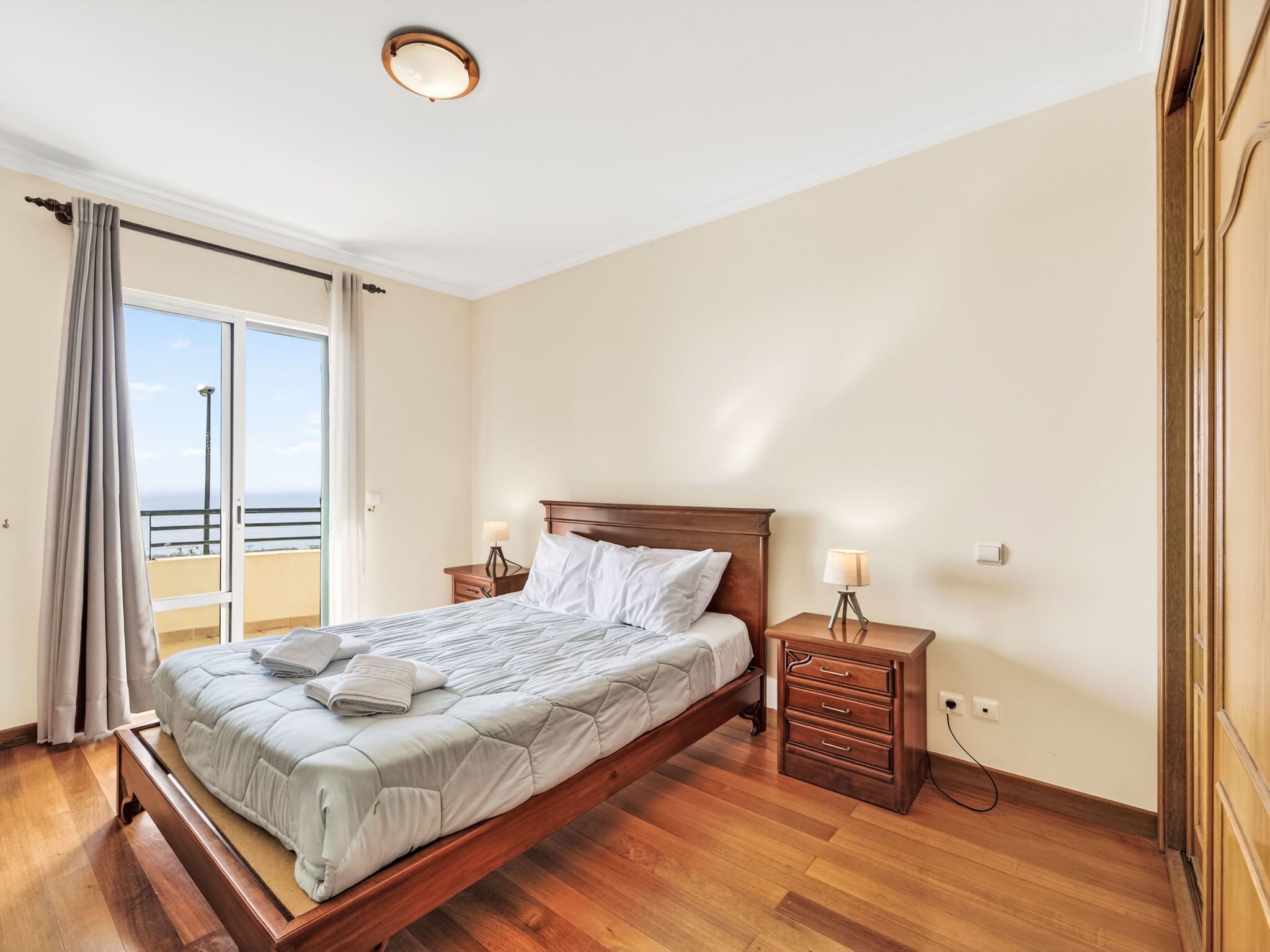 A bedroom with a wooden bed, a nightstand, a drawer unit, and a view of the sea through a sliding door.
