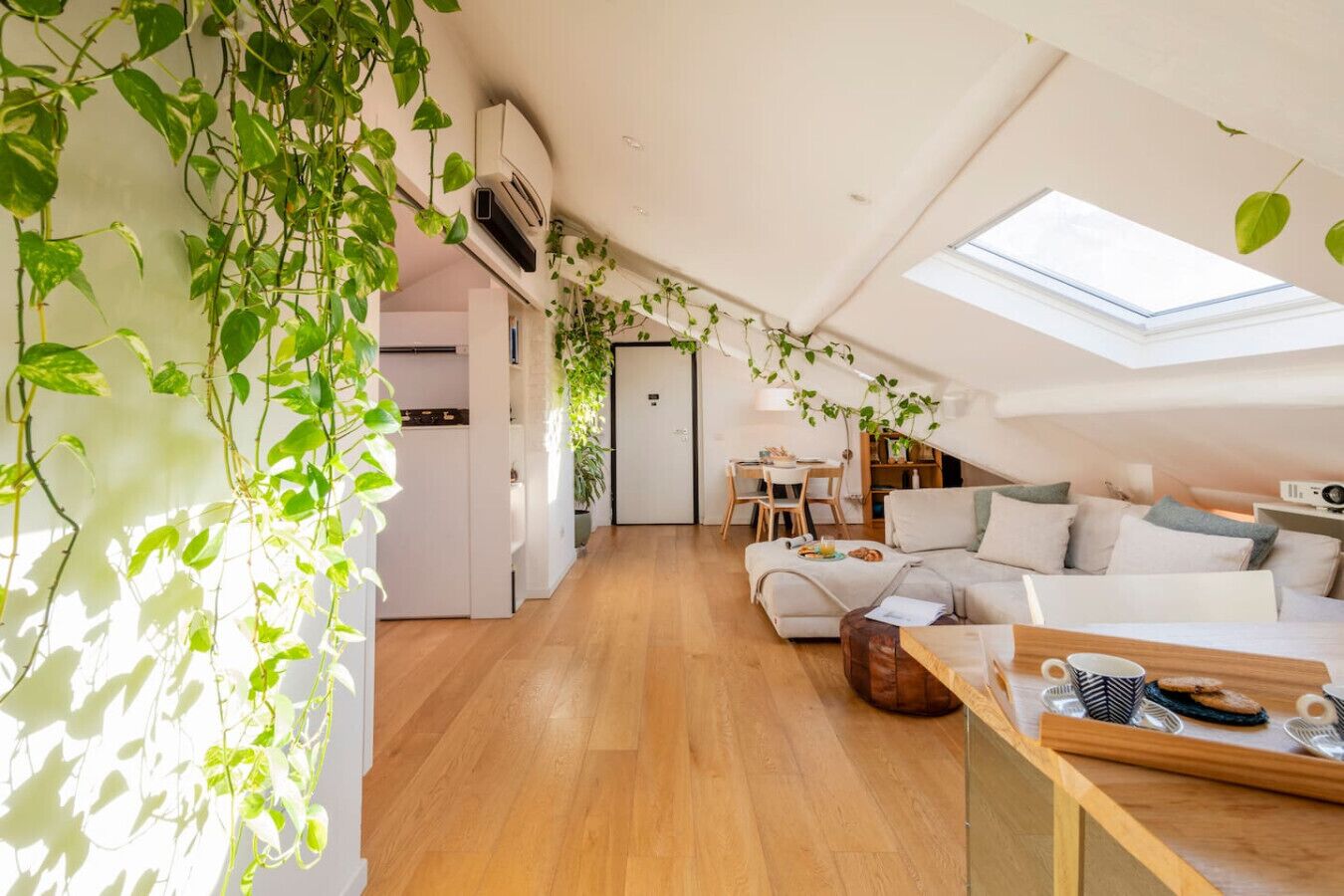 A modern loft apartment with a skylight, wooden flooring, a sofa, a dining area, and green plants.