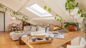 En hyggelig loftlejlighed med ovenlys, spiseplads, sofa, sofabord med morgenmadsprodukter og forskellige planter.