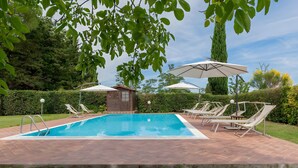 Una piscina rettangolare con bordo piastrellato, circondata da lettini e ombrelloni, con alberi e un siepe sullo sfondo.