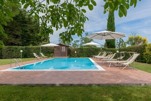 Ein rechteckiger Pool mit gefliestem Sonnendeck, umgeben von Liegestühlen und Sonnenschirmen, vor einer Kulisse aus Bäumen und einer Hecke.