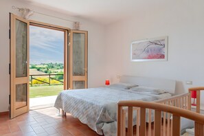 Ein Schlafzimmer mit einem Bett, Blick durch eine offene Tür in einen Garten, einem Bild an der Wand und einer roten Lampe.