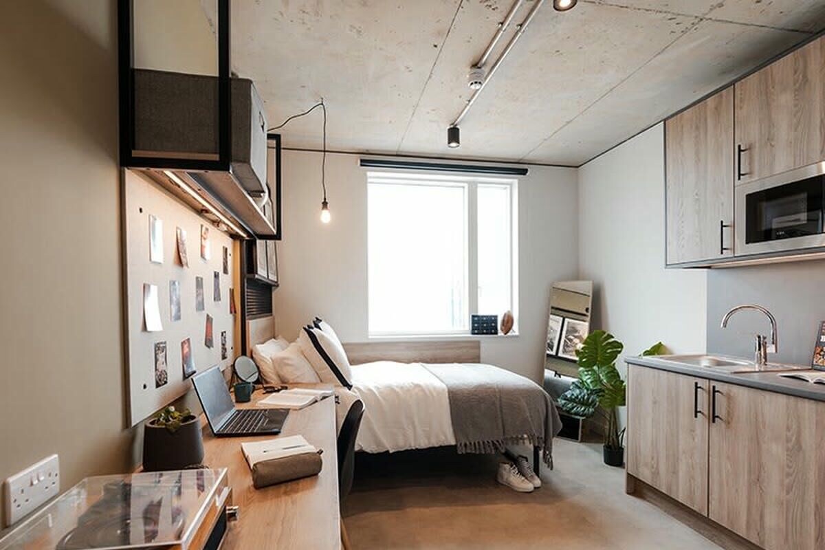 Ein modernes Schlafzimmer mit einem Bett, einem Schreibtisch mit einem Laptop, einem Fenster und einer Küchenzeile.