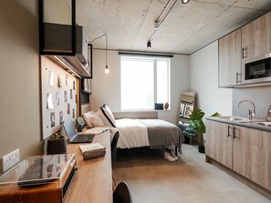Ein modernes Schlafzimmer mit einem Bett, einem Schreibtisch mit einem Laptop, einem Fenster und einer Küchenzeile.