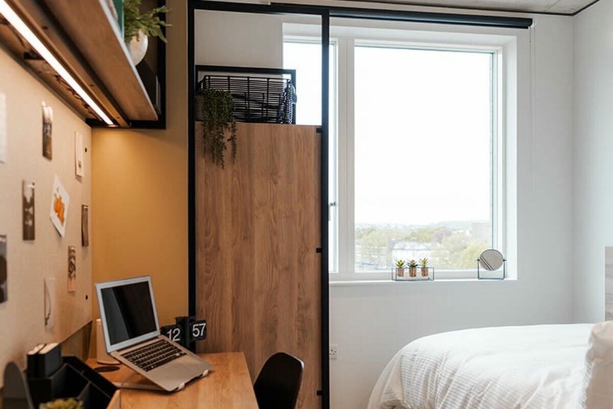Ein Schlafzimmer mit Bett, Fenster mit Blick, Schreibtisch mit Laptop und Tür mit Nummer.