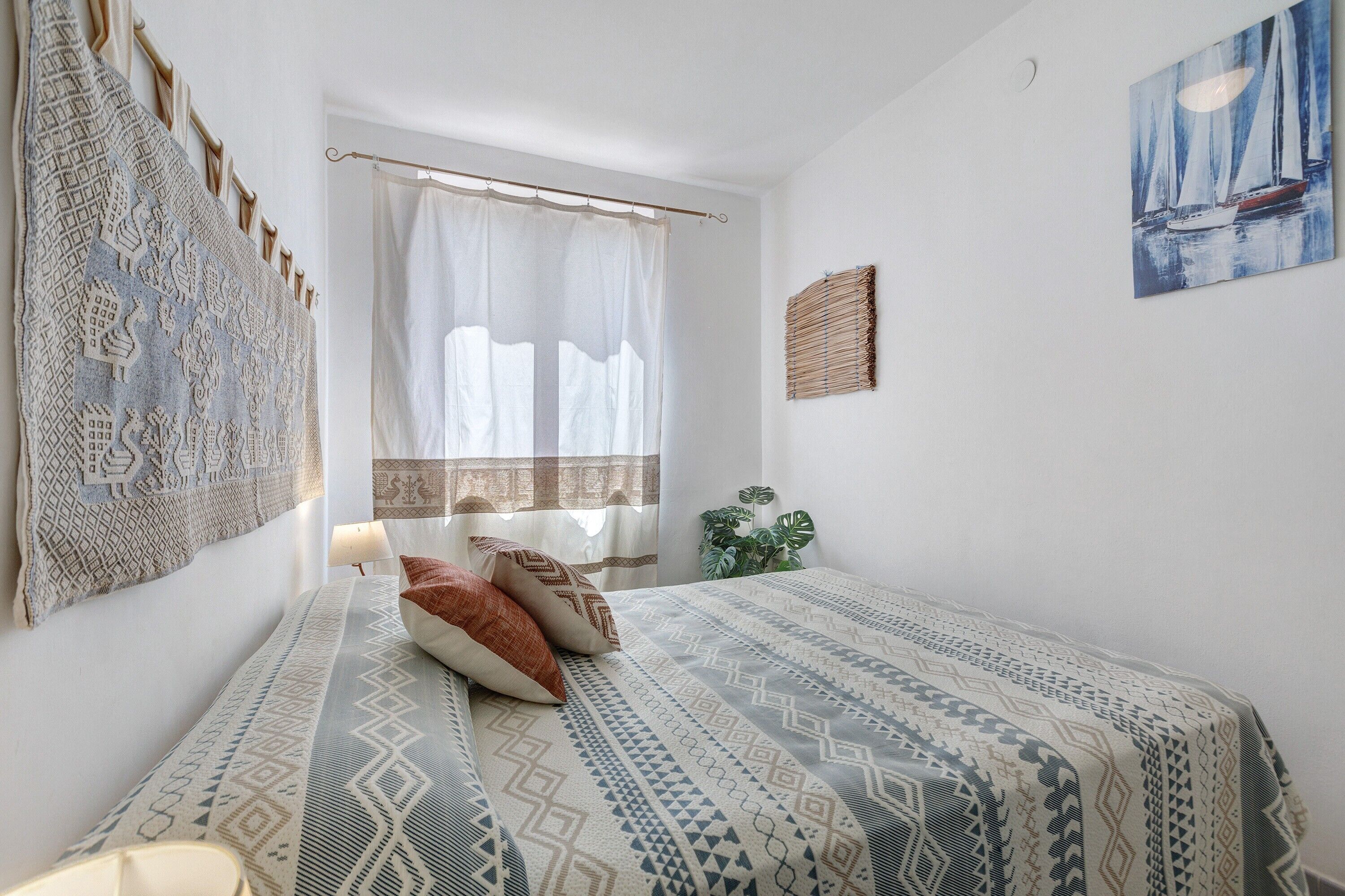 Une chambre à coucher avec un lit, une fenêtre ornée de rideaux légers, une œuvre d’art accrochée au mur et une plante en pot.